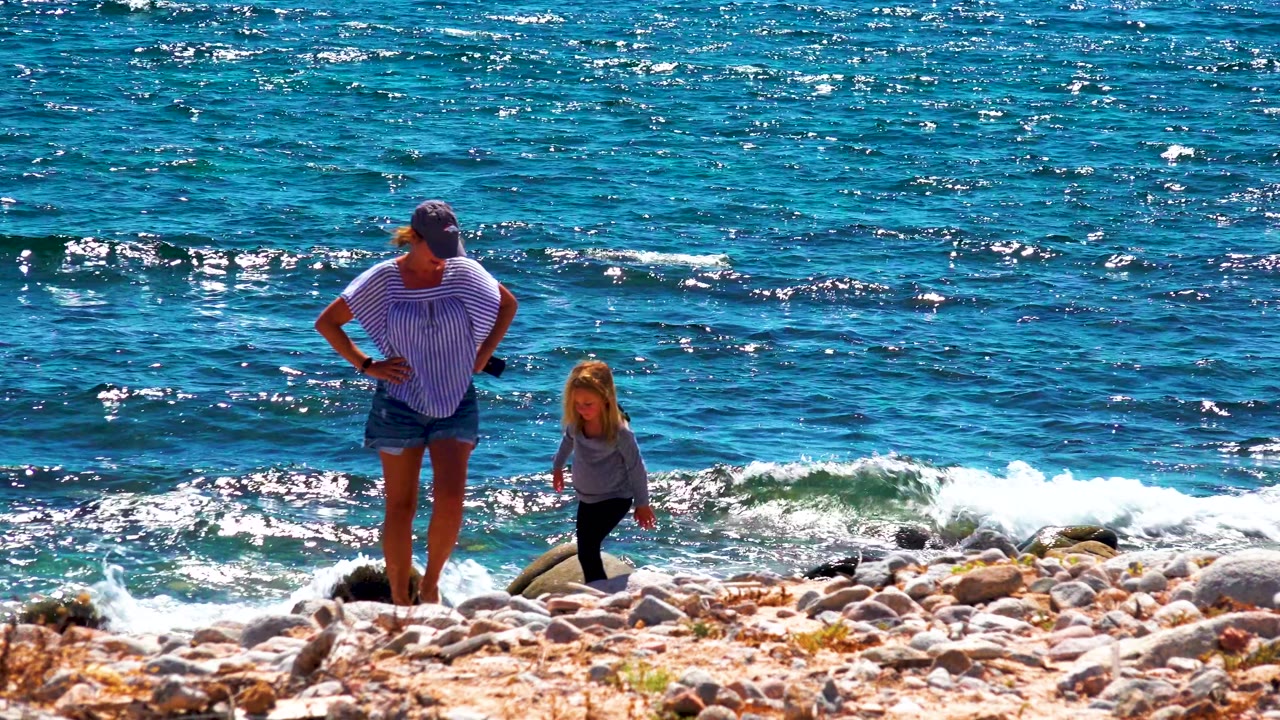 Adult stands near a child on rocks as waves break behind them.