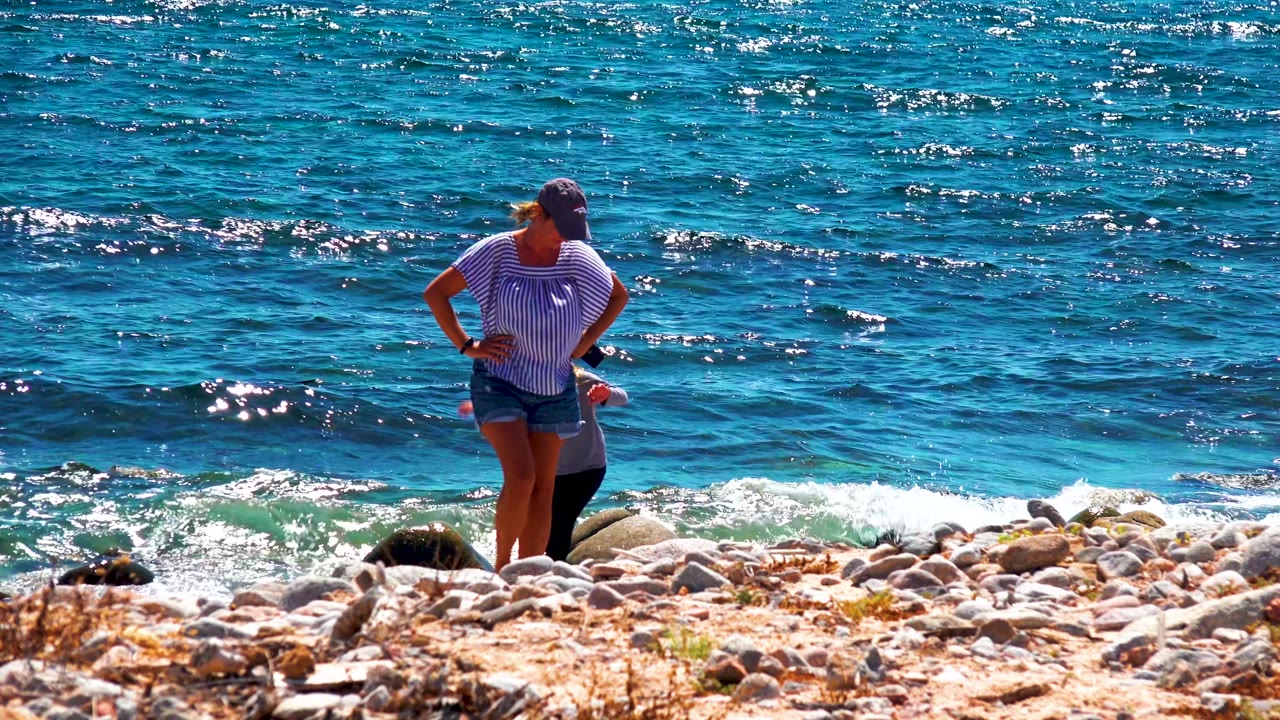 At El Sargento La Ventana Mexico, the adult stands on the rocks while the child is partly visible near the shoreline and choppy blue water fills the background.