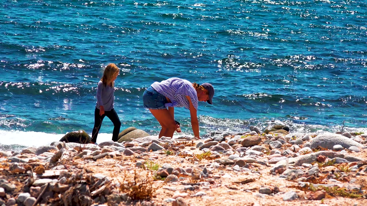 In El Sargento La Ventana Mexico, an adult bends toward the rocky beach while a child walks past at the edge of bright blue ocean water.
