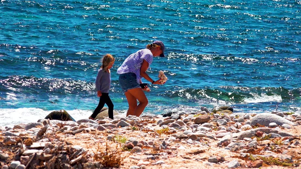 Adult stands with an object while a child approaches along a rocky ocean shore.