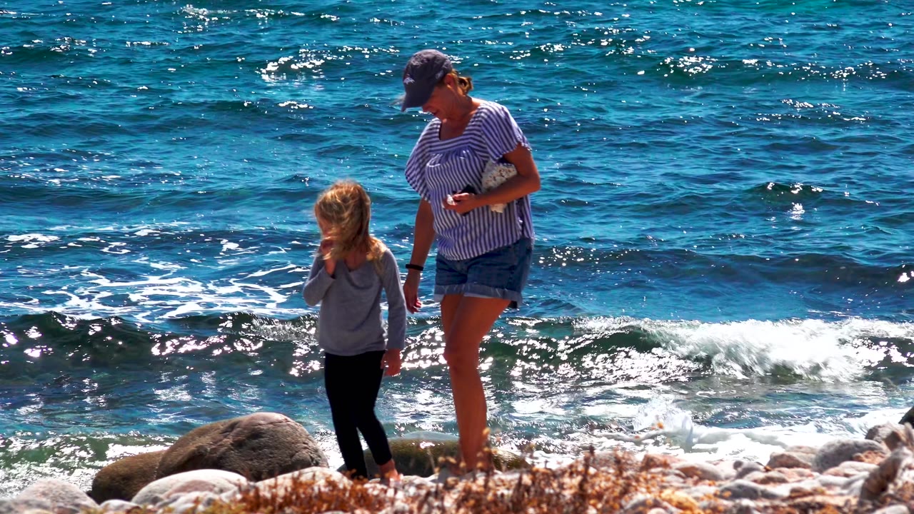 At El Sargento La Ventana Mexico, a woman in a striped shirt stands near a young girl on a rocky shore as blue waves break behind them.