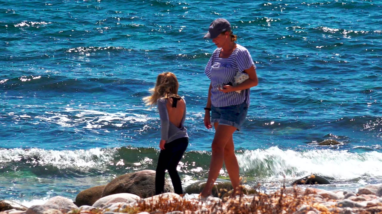 Young girl and woman face each other on rocks near breaking surf.