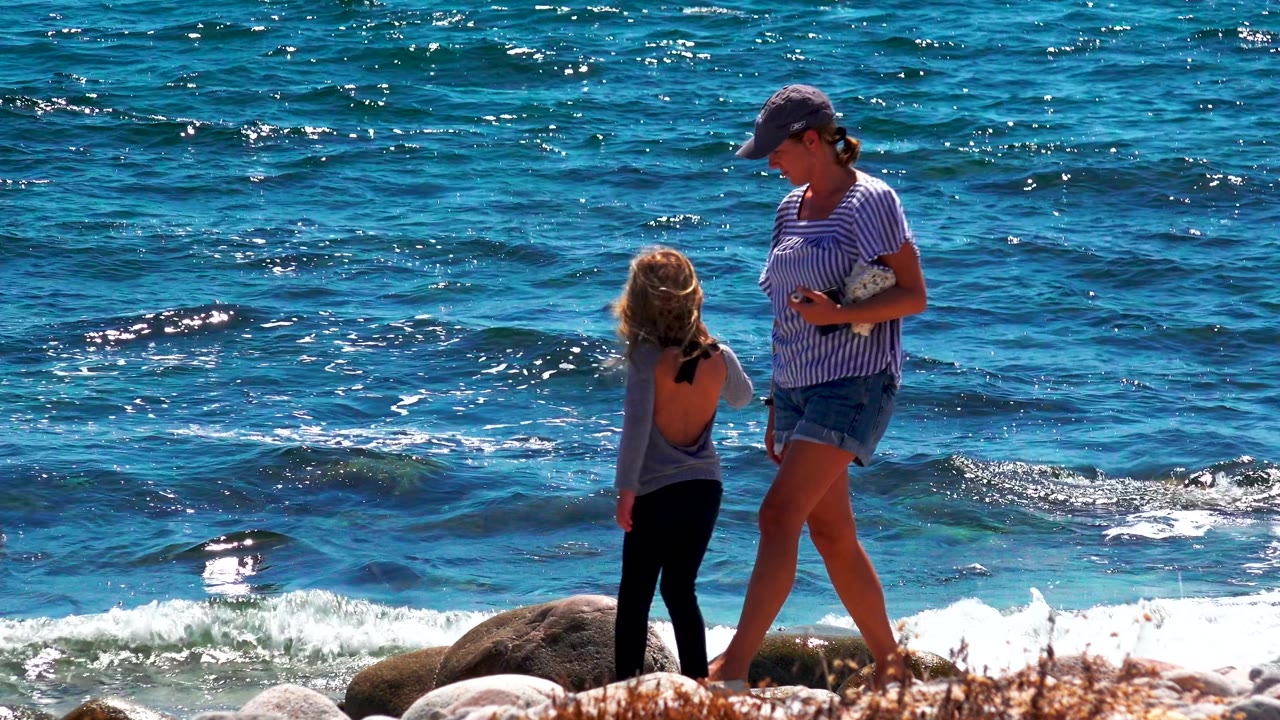 At El Sargento La Ventana Mexico, the woman steps along the rocks beside the young girl as darker blue water and small white wave crests fill the background.