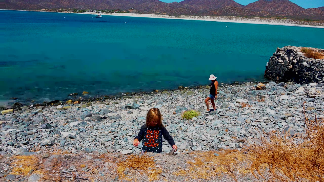 At El Sargento La Ventana Mexico, a child sits on the rocky slope above the shoreline while an adult in a white hat stands near the clear blue water and mountains line the background.