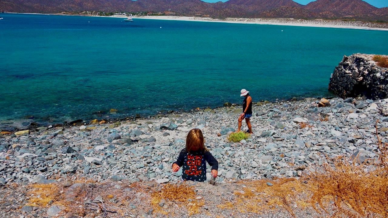 Child seated on pebbles with an adult walking along the rocky shoreline.