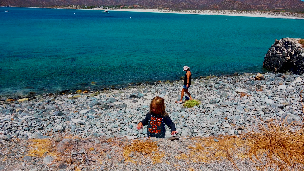 At El Sargento La Ventana Mexico, the child sits facing the shoreline as the adult stands farther right near the water, with turquoise sea, pebbles, dry grass, and mountains visible.