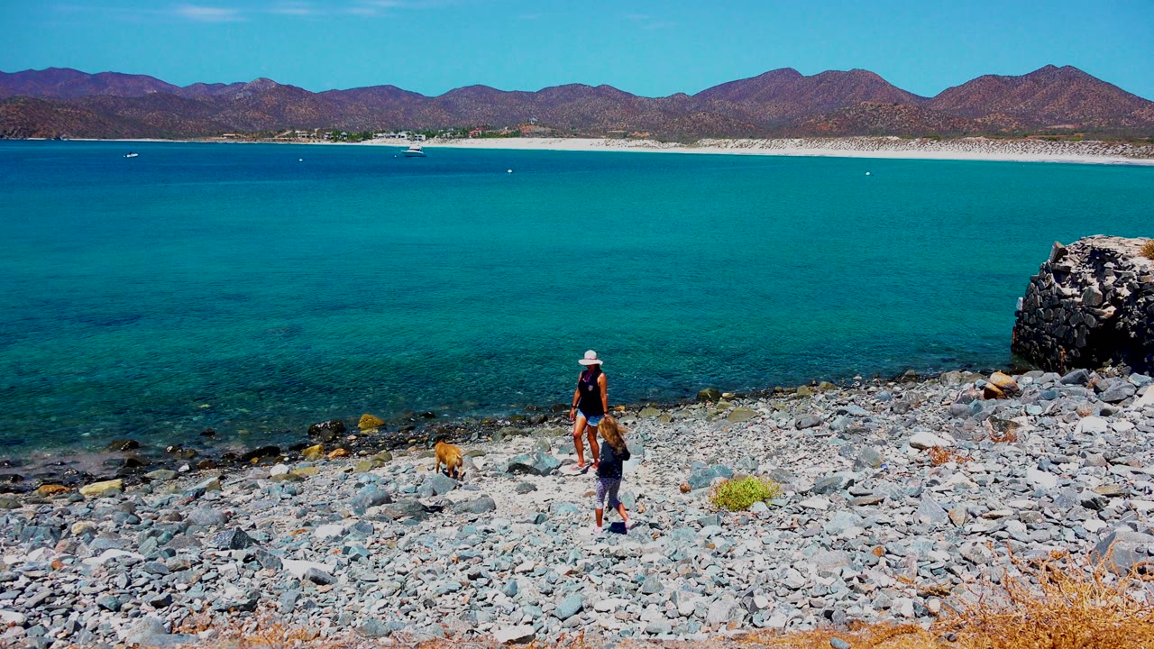 At 0:00 in El Sargento La Ventana Mexico, a hat-wearing person stands with a smaller figure and brown dog on gray rocks below turquoise water and distant mountains.