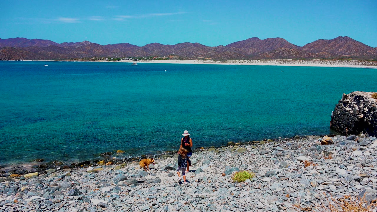 At 0:02 in El Sargento La Ventana Mexico, the person and dog appear near the center-right of the rocks while the broad turquoise bay and brown mountains stay level behind them.
