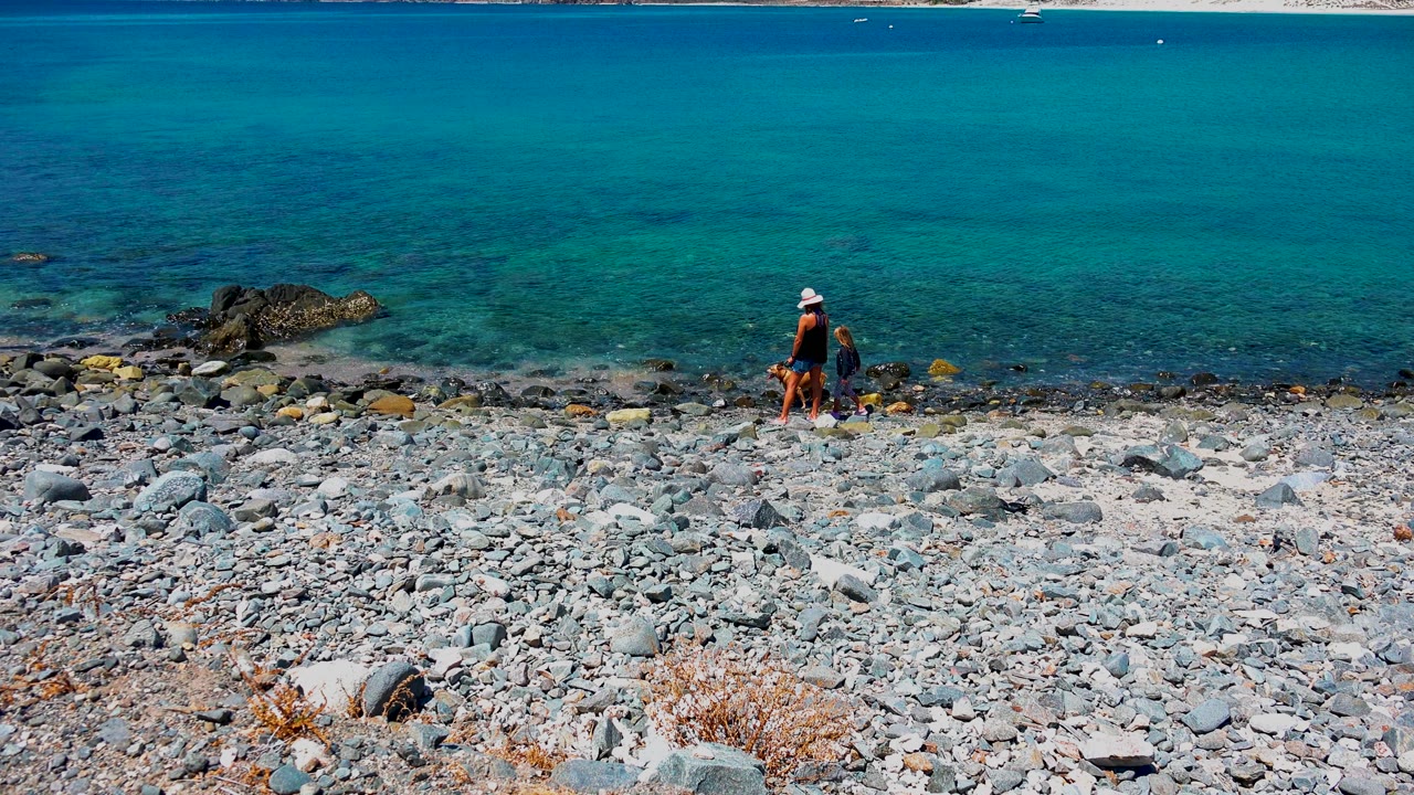 At 0:00 in El Sargento La Ventana Mexico, an adult in a light hat and a child walk along a pebbly shore with turquoise water and distant mountains behind them.