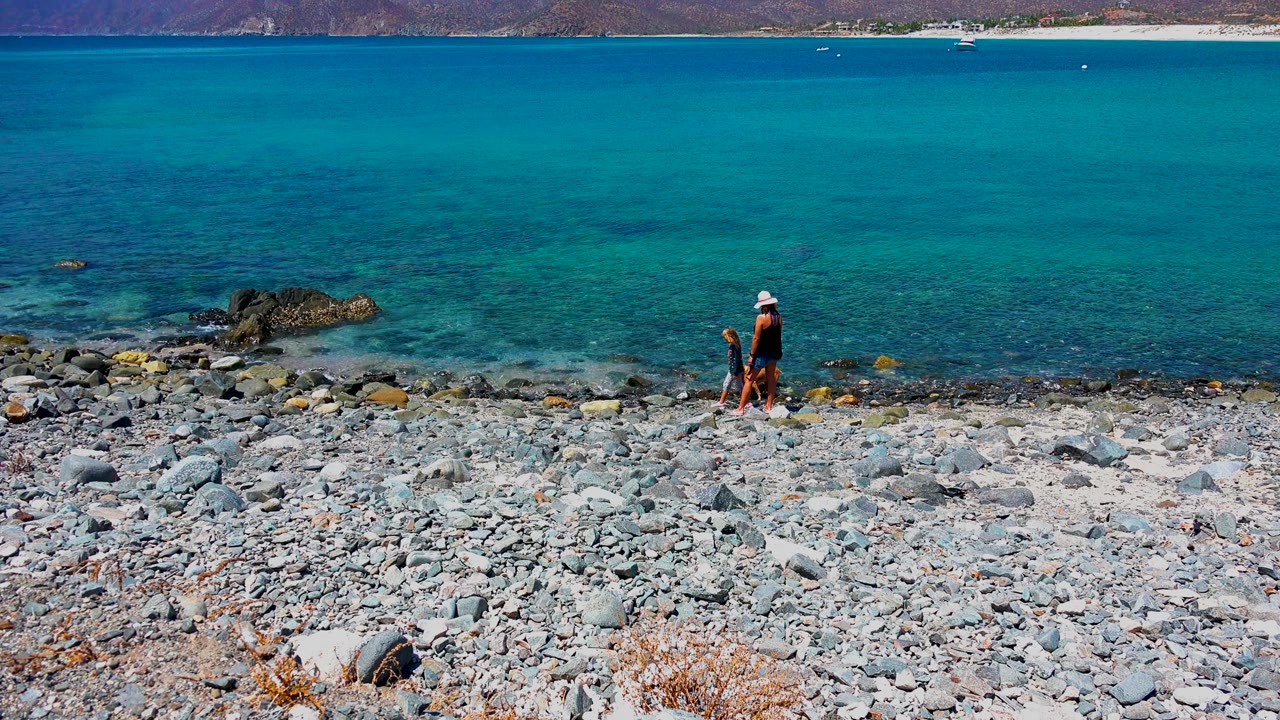 At 0:02 in El Sargento La Ventana Mexico, the adult and child appear near the right side of the wide frame, with pale beach stones in front and clear blue water stretching to distant mountains.