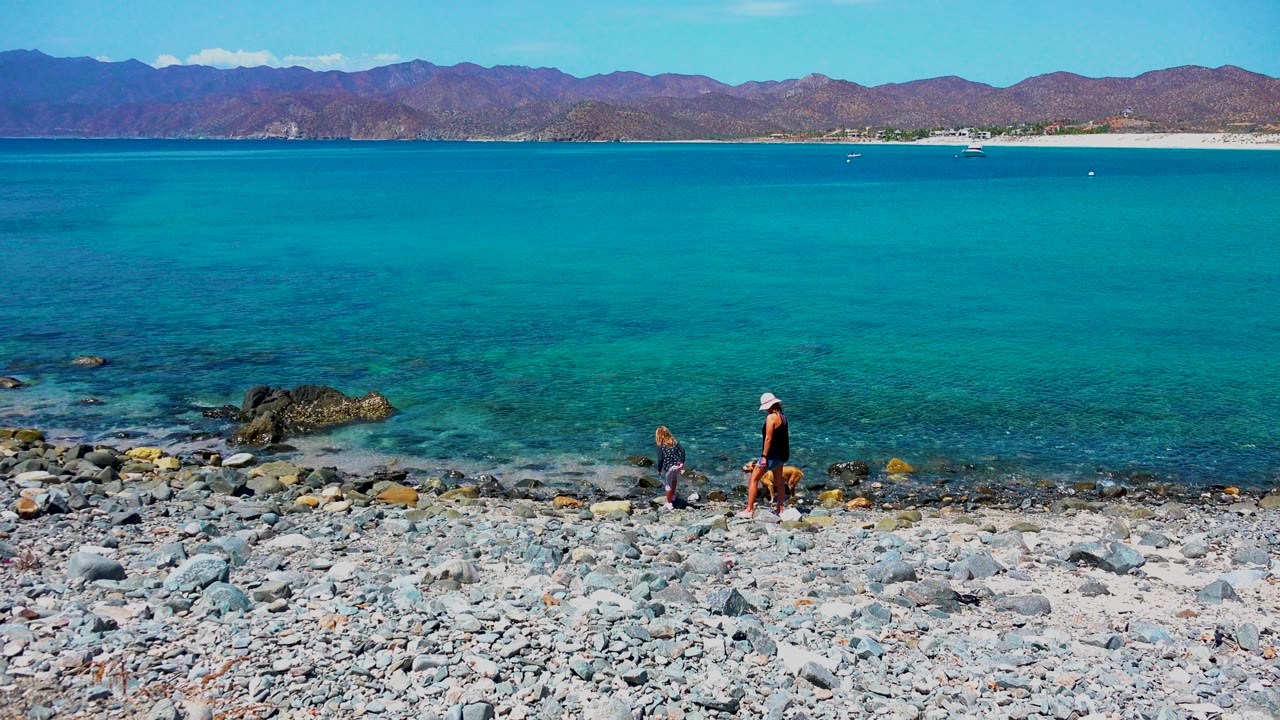 At El Sargento La Ventana Mexico, a person in a white cap and a brown dog walk along a rocky pebble beach with turquoise water and mountains behind them.