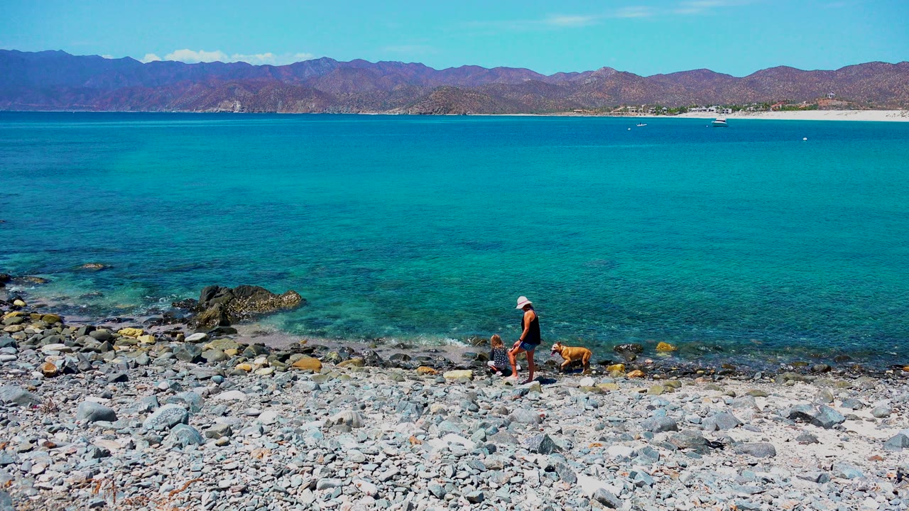 At El Sargento La Ventana Mexico, a beachgoer and brown dog move near the right side of the pebble beach, with dark rocks in the shallow turquoise water.