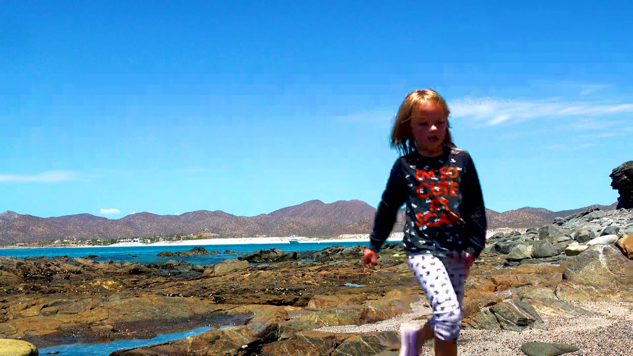 At 0:00 in El Sargento La Ventana Mexico, a young child walks toward camera across rocks and gravel with blue ocean and low mountains in the background.