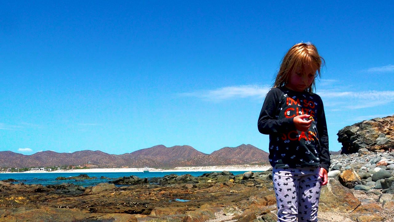 At 0:02 in El Sargento La Ventana Mexico, the child stands near the right side of frame beside large rocks, with bright blue water and brown mountains behind her.
