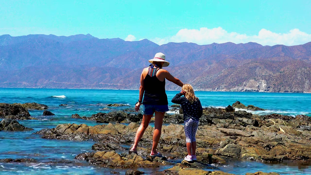 At El Sargento La Ventana Mexico, a woman in a sun hat and a child face the rocky shoreline with turquoise water and distant mountains behind them.