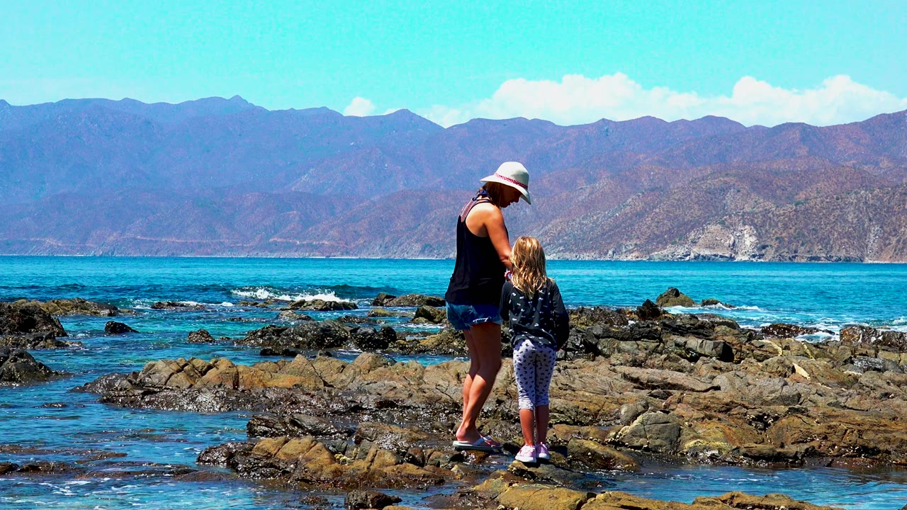 At El Sargento La Ventana Mexico, the woman and child look down toward their footing on the rocks as blue water and brown mountains fill the background.