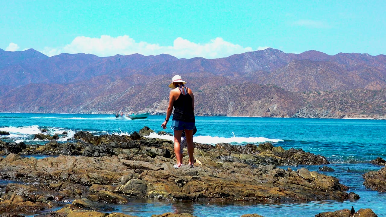 At El Sargento La Ventana Mexico, a woman in a white hat stands on dark rocks facing turquoise ocean, with a small boat offshore and mountains across the horizon.