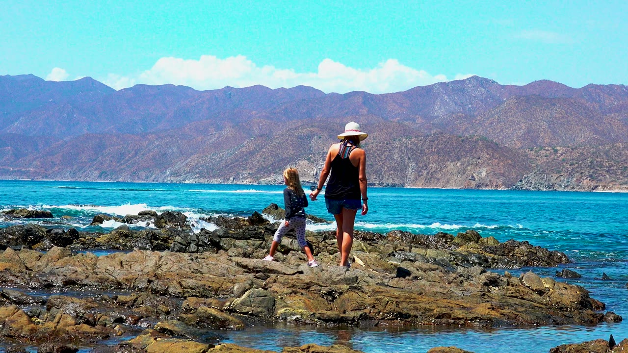 Child walks toward woman on rocks beside turquoise water and distant mountains.