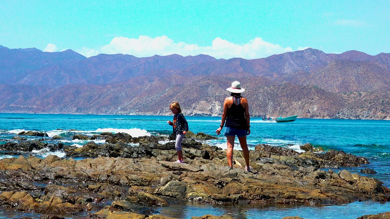 At El Sargento La Ventana Mexico, the woman and child stand on separate sections of rock while a small boat sits offshore against the blue water and mountain backdrop.