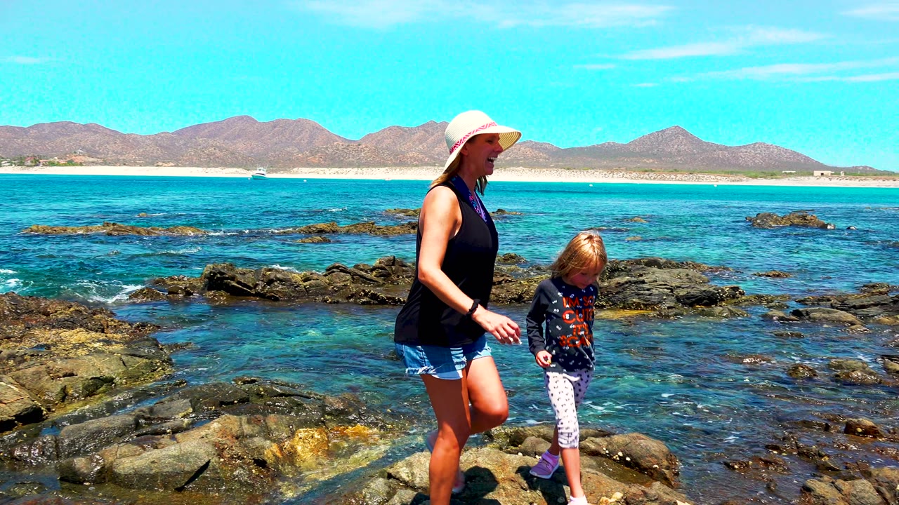 At 0:00 in El Sargento La Ventana Mexico, a woman in a sun hat and a young child walk across dark beach rocks with blue water, pale sand, and mountains behind them.