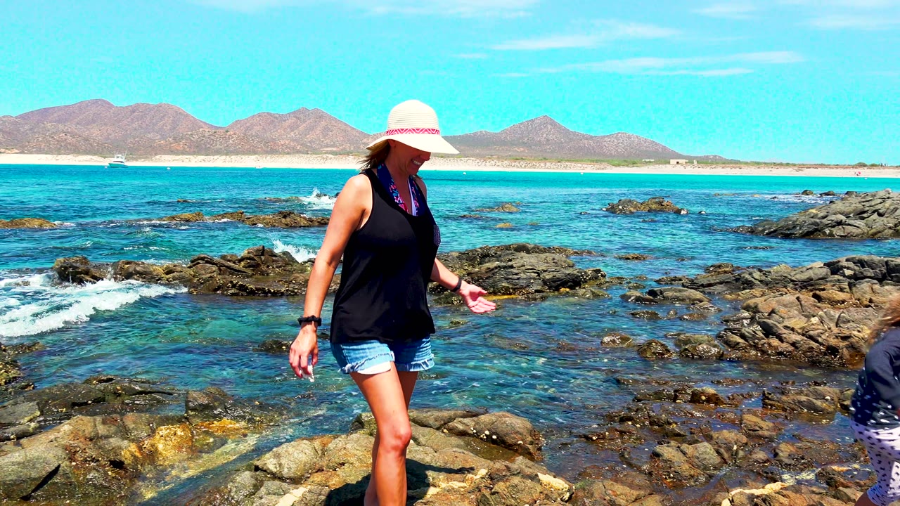At 0:02 in El Sargento La Ventana Mexico, the woman walks to the right across uneven rocks while the young child is only partly visible at the far right edge.