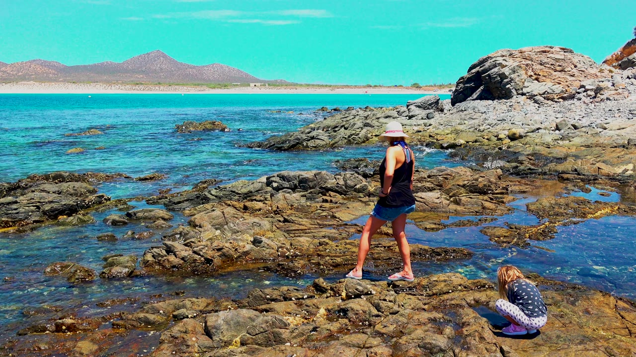 At 0:00 in El Sargento La Ventana Mexico, a woman stands on wet shoreline rocks while a child crouches near the lower right, with clear blue water and distant hills behind them.