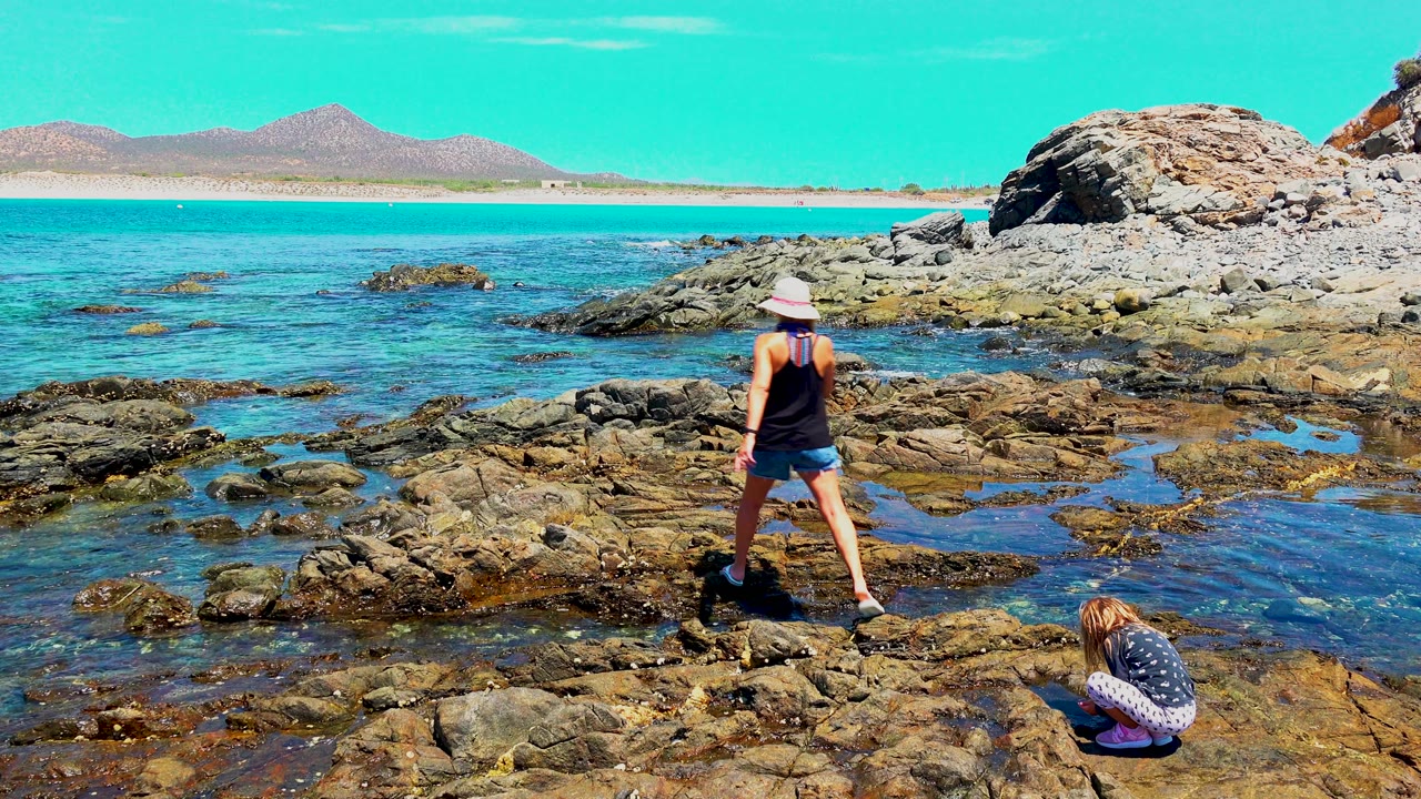 At 0:02 in El Sargento La Ventana Mexico, the woman stands with her feet apart on uneven wet rocks while the child crouches in the lower right foreground near reflective tide pools.