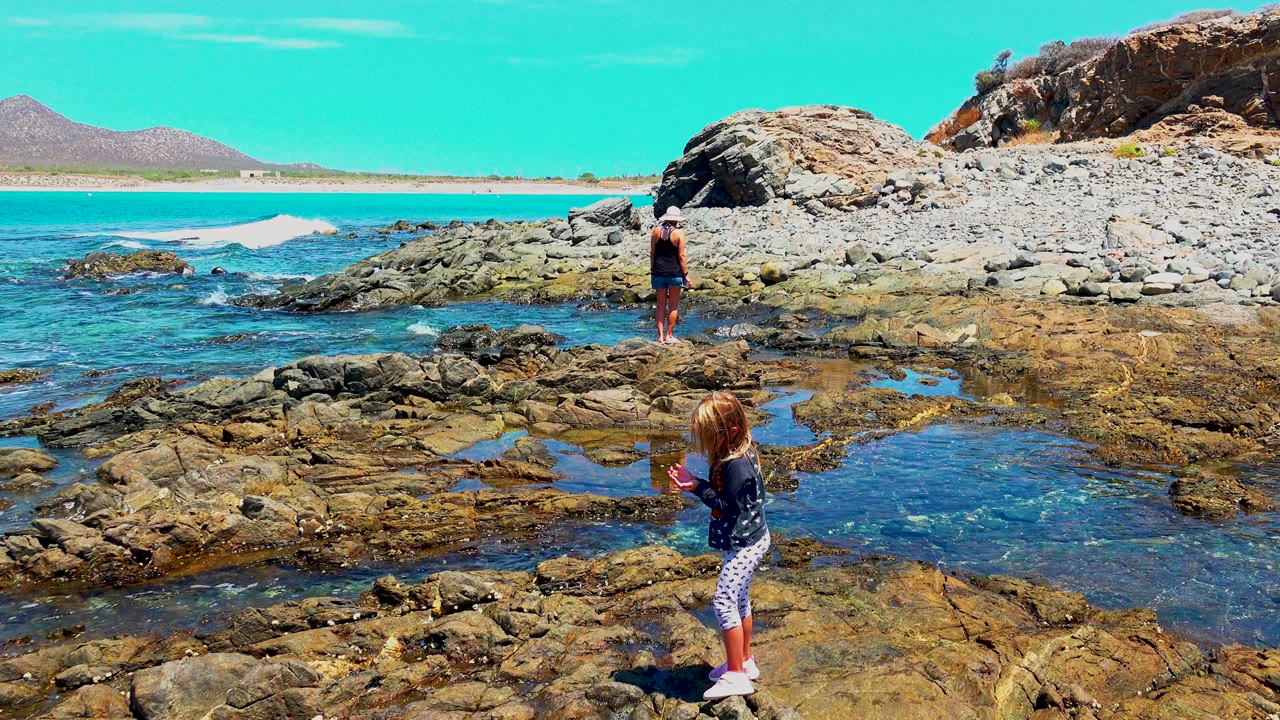 At El Sargento La Ventana Mexico, two children stand apart on rocky tide pools, with clear blue water in the foreground and a sandy beach and low hills in the distance.