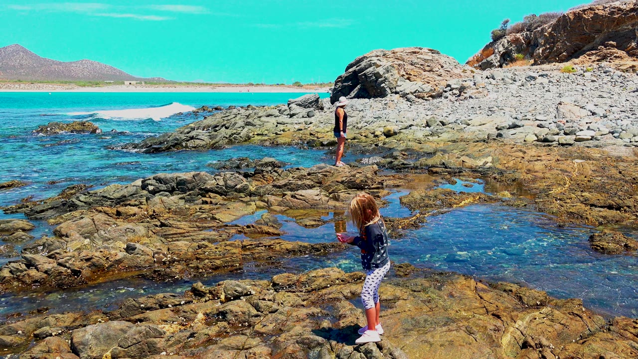 Child in foreground near tide pool with another child on rocks.