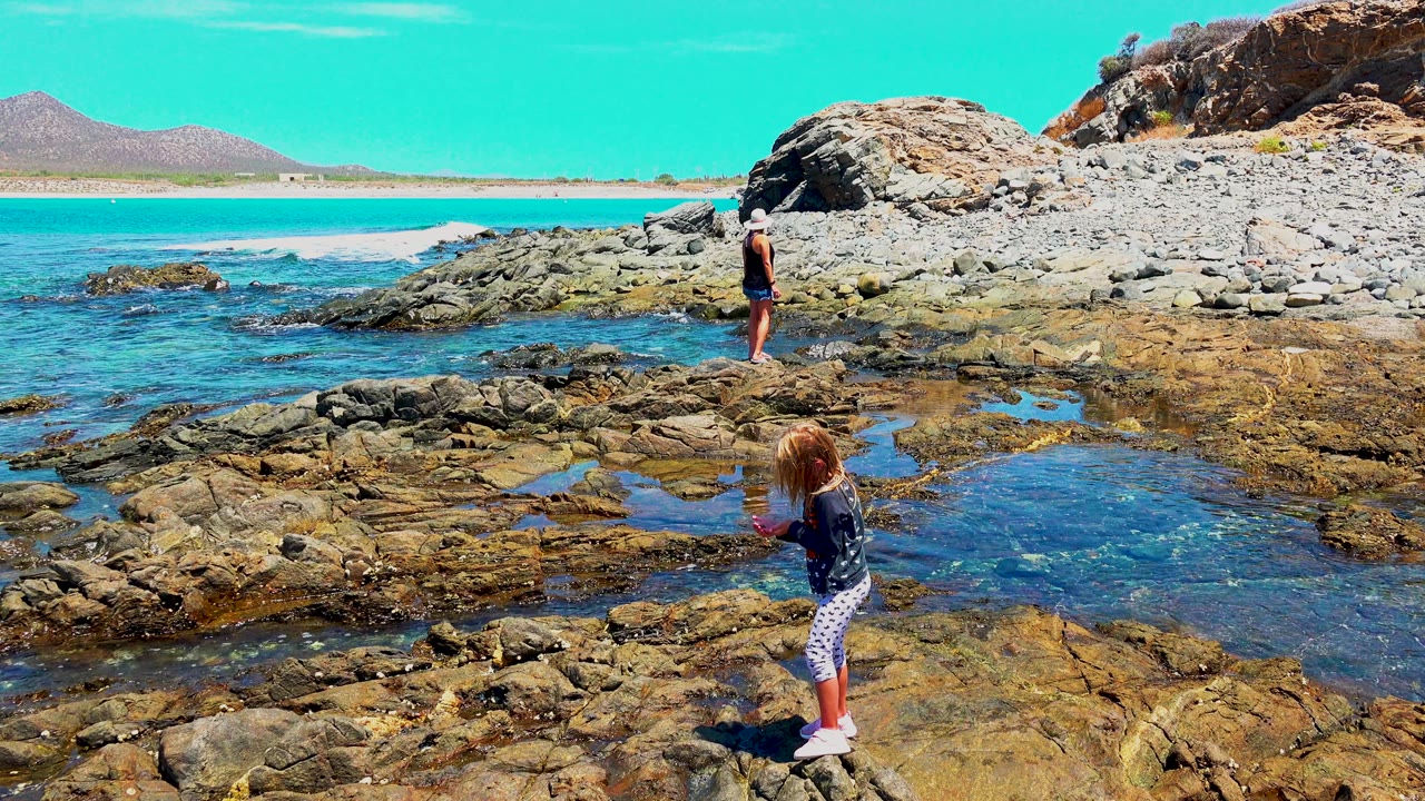 At El Sargento La Ventana Mexico, two children remain on the sunlit rock shelf, with rippled tide-pool water in front and turquoise ocean and pale sand behind.