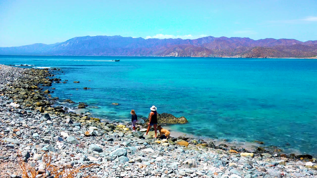 At 0:00 in El Sargento La Ventana Mexico, two people and a dog stand on a rocky shore beside clear blue water, with a small boat offshore and mountains across the horizon.