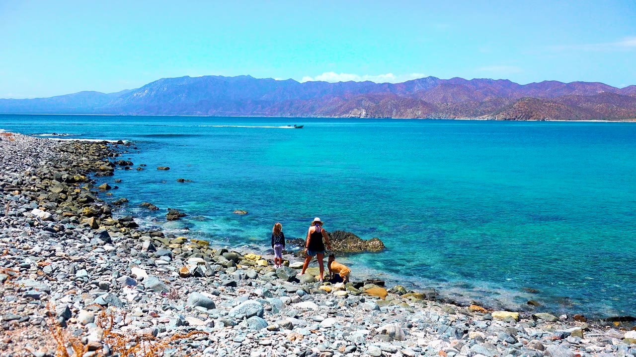 At 0:02 in El Sargento La Ventana Mexico, two people and a dog are positioned near dark shoreline rocks, with turquoise shallows, deeper blue water, a boat, and mountains behind them.