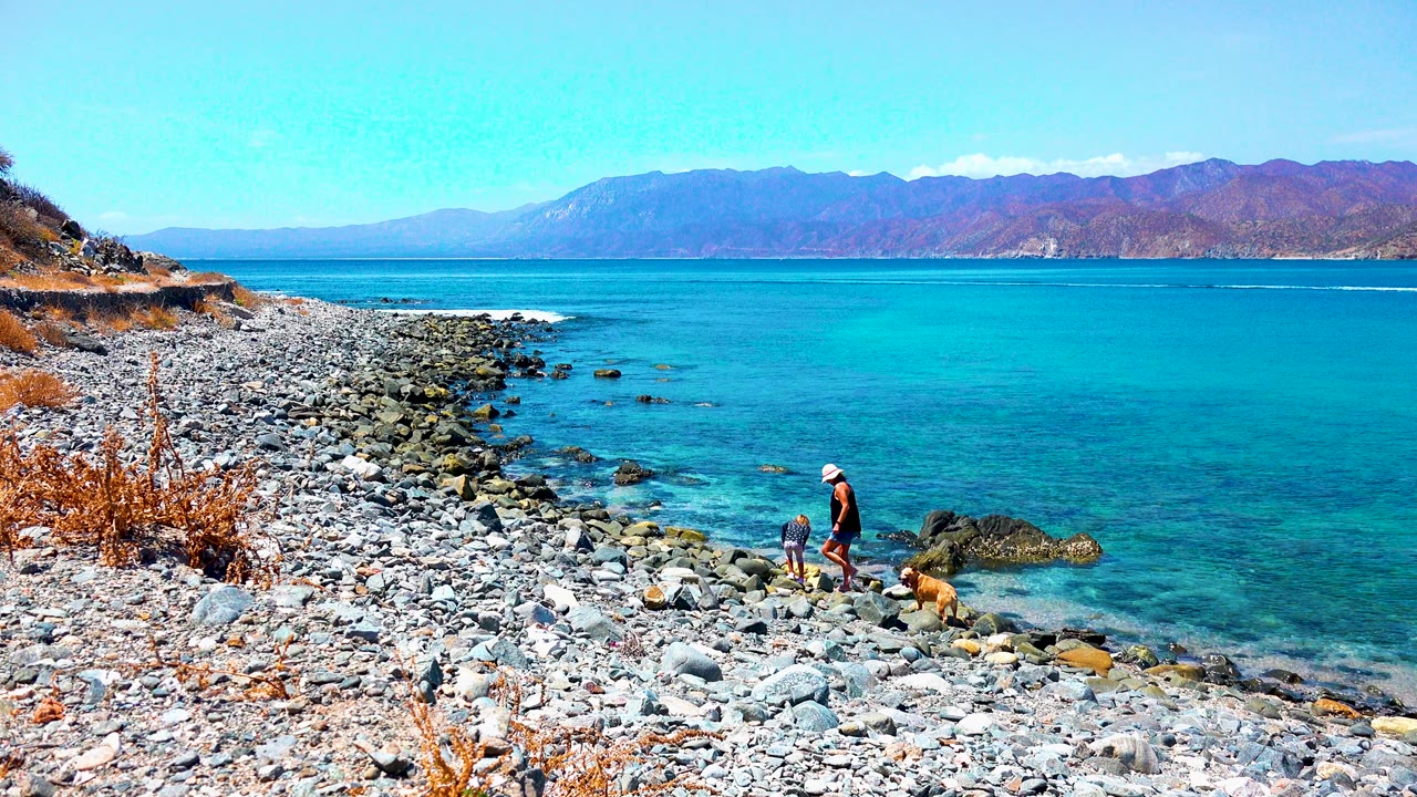 At El Sargento La Ventana, Mexico, a person and dog stand near the waterline on a pebble-covered shore with turquoise ocean and mountains in the background.