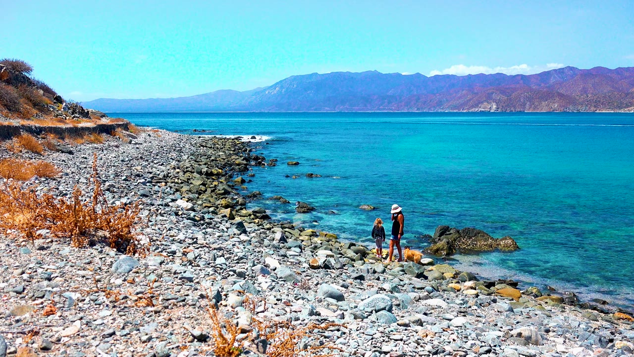 At El Sargento La Ventana, Mexico, two people stand together on the rocky beach while a dog moves near the waterline and mountains run across the horizon.