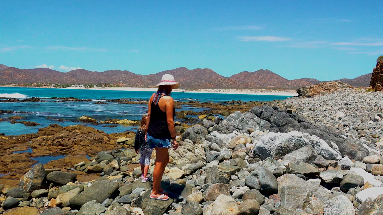 At 0:00 in El Sargento La Ventana, Mexico, a woman in a white hat and a child stand on large gray rocks with tide pools, blue water, surf, and brown mountains behind them.