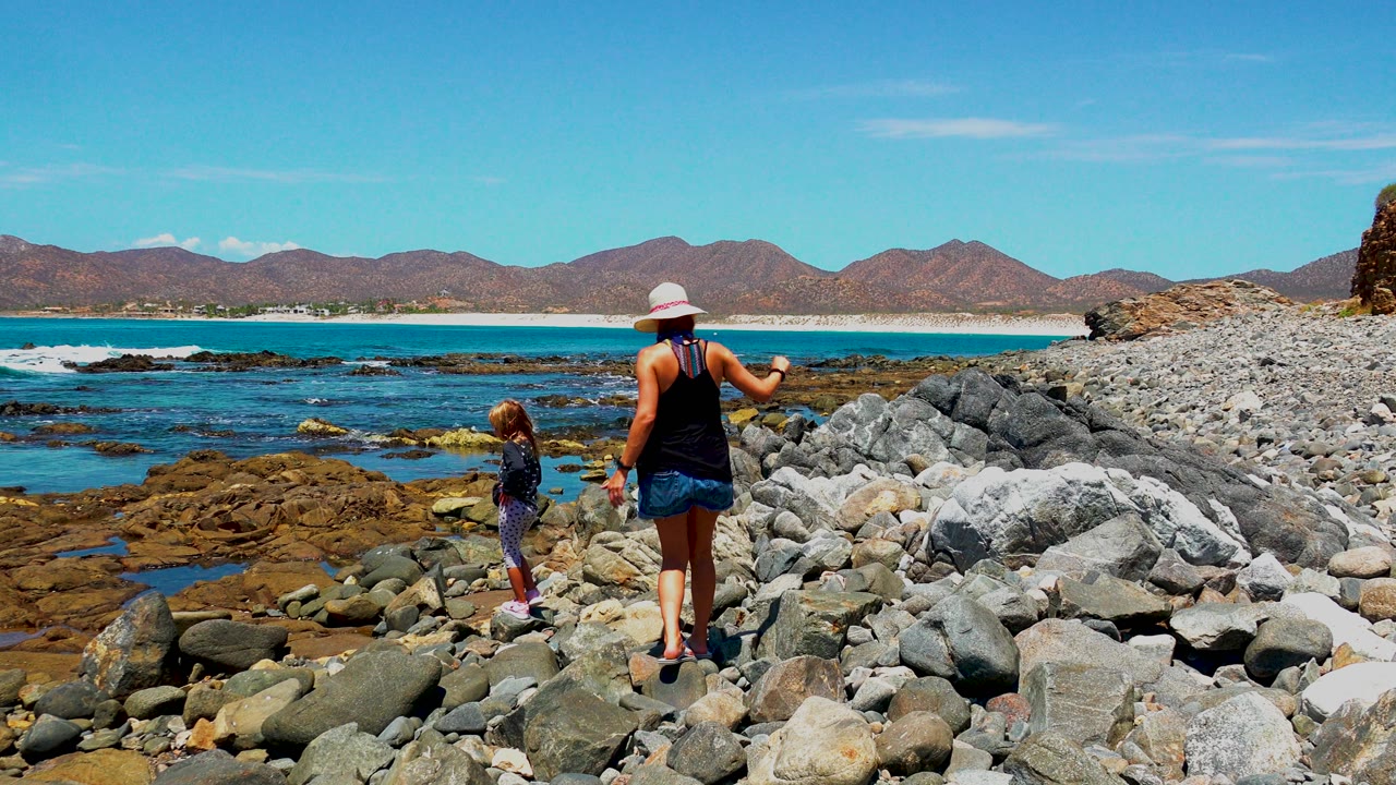 At 0:02 in El Sargento La Ventana, Mexico, the woman and child are separated across the rocky foreground, with the child near the tide pools and the woman turned toward the shoreline.