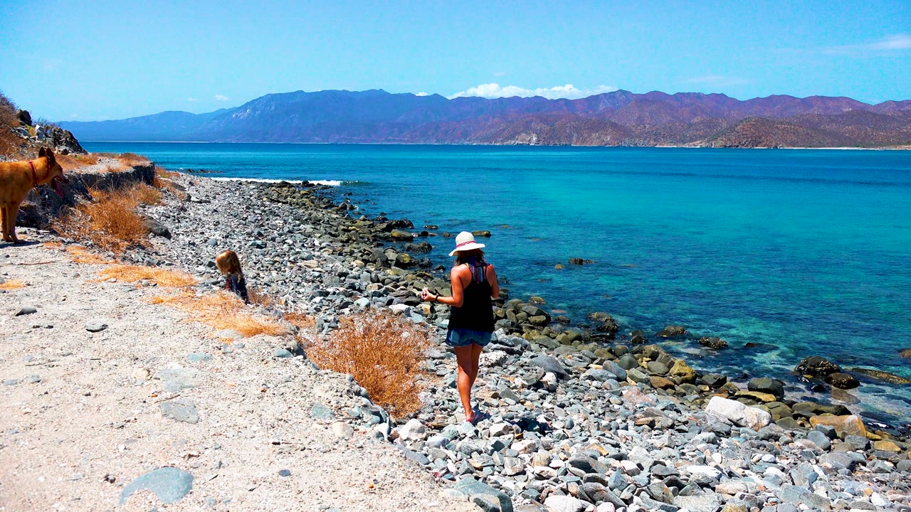 In El Sargento La Ventana Mexico, the woman pauses near the waterline while the child crouches among the shore rocks and the dog stands on the higher path at left.