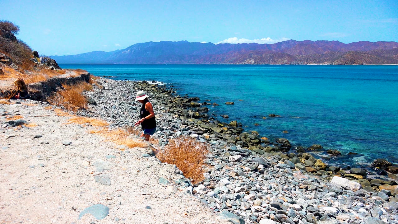 At El Sargento La Ventana, Mexico, a person in a white hat stands near a rocky shore with turquoise water to the right and mountains across the horizon.
