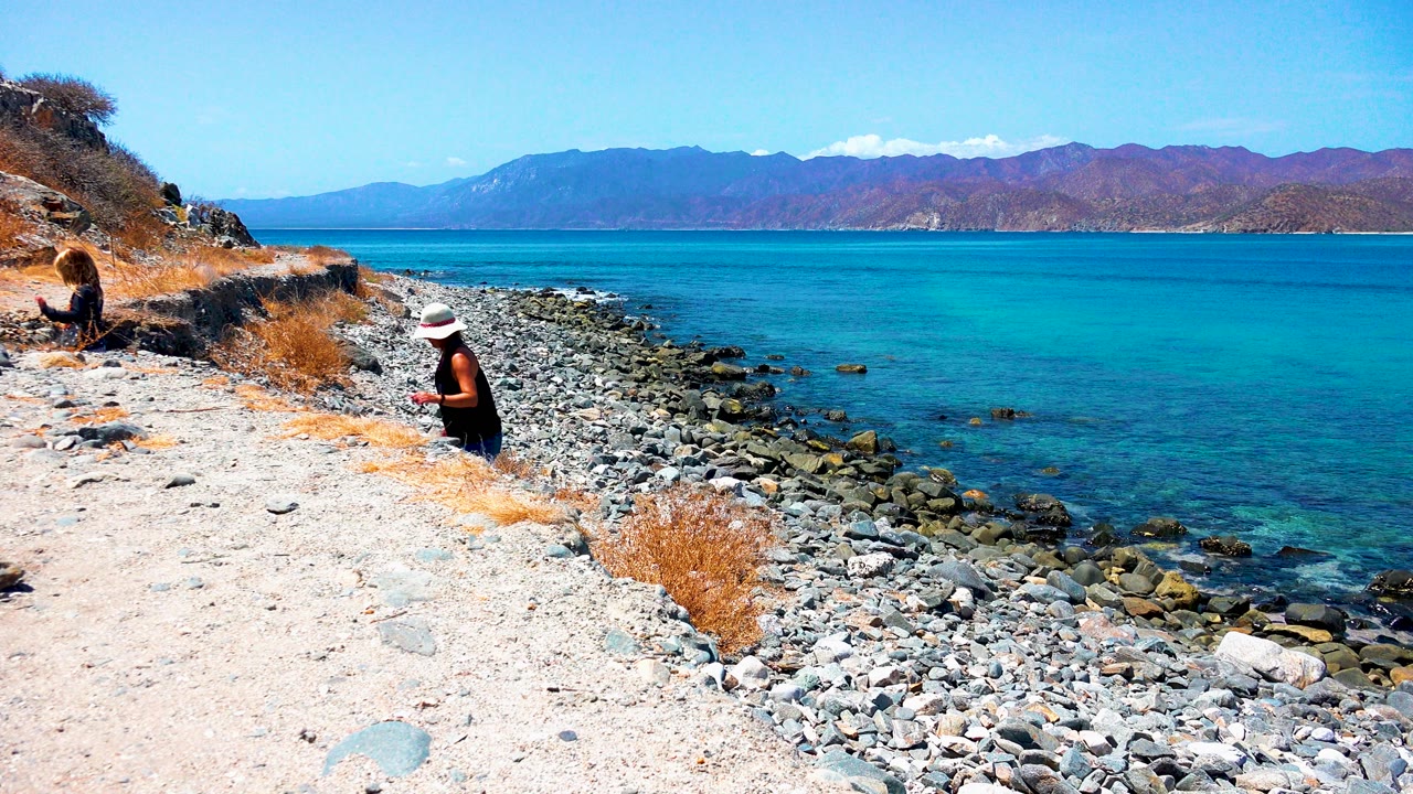 Person in a white hat crouches near rocks on a dry coastal path above blue water.