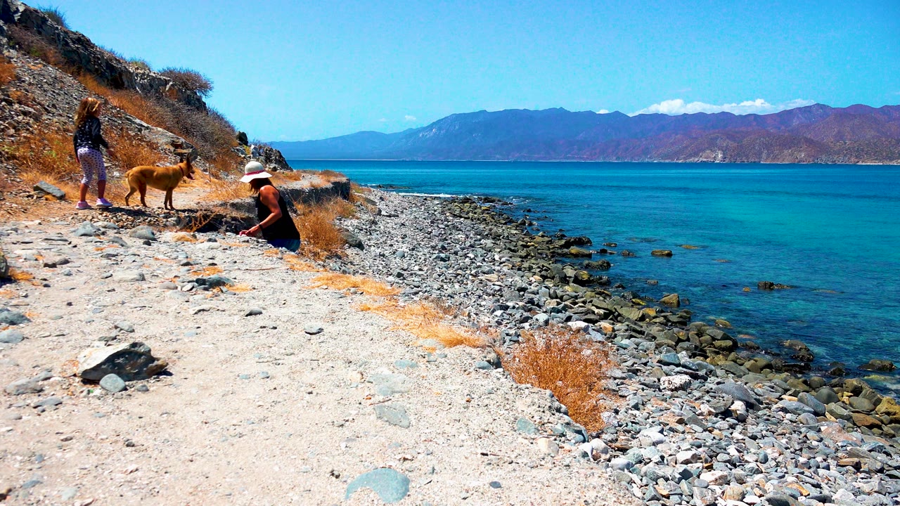At 0:00 in El Sargento La Ventana Mexico, two people and a tan dog occupy the left side of a rocky shore while clear blue water and distant mountains fill the right background.