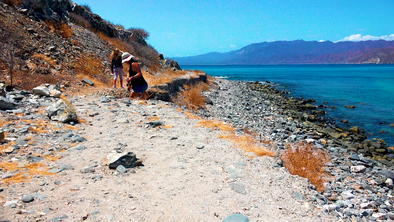 In El Sargento La Ventana Mexico, a woman bends near the rocky coastal path while a child stands nearby, with blue ocean and mountains beyond.