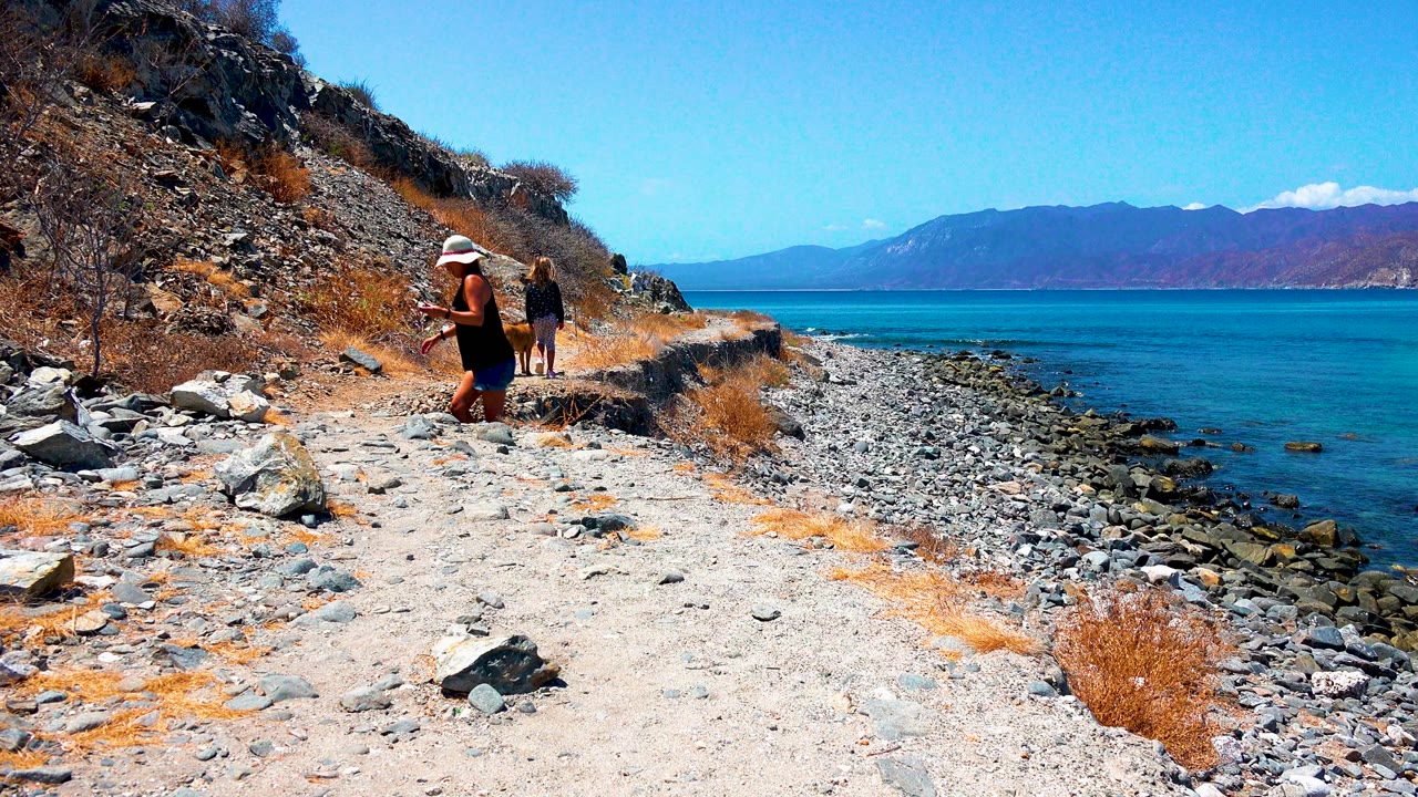 In El Sargento La Ventana Mexico, the woman and child continue away along the coastal path, with loose stones, dry hillside brush, blue water, and a mountain horizon visible.
