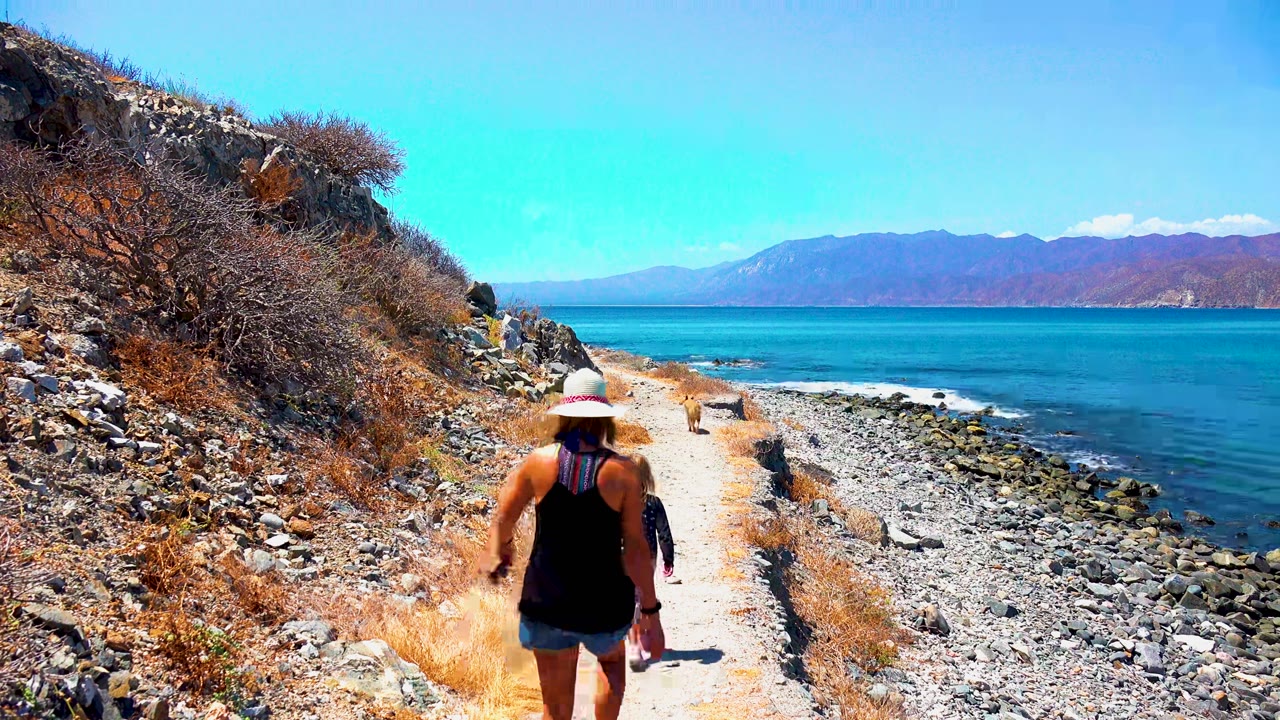 At 0:00 in El Sargento La Ventana Mexico, a woman in a sun hat walks away on a pale rocky trail with dry brush on the left and blue ocean to the right.