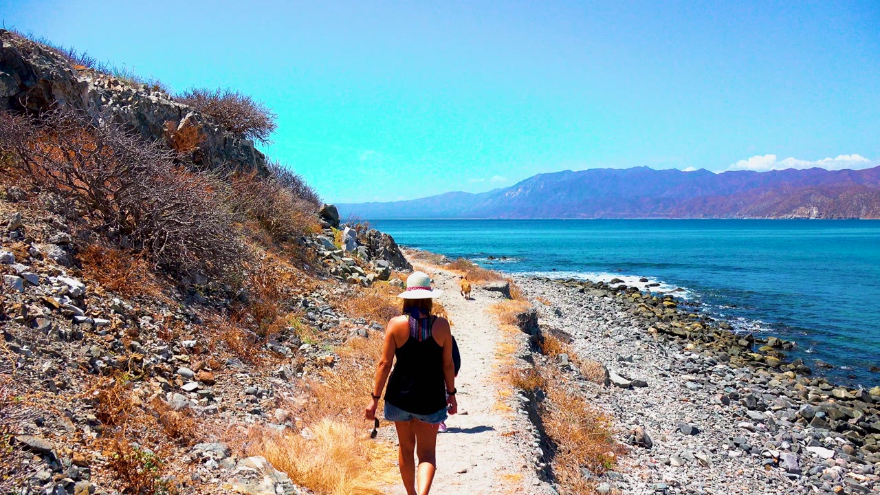 Woman walking farther down a dry coastal path with ocean and mountains beyond.