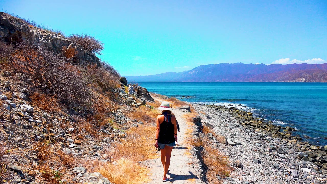 At 0:02 in El Sargento La Ventana Mexico, the woman walks away while a child becomes visible behind her on the rocky coastal trail beside the blue sea.