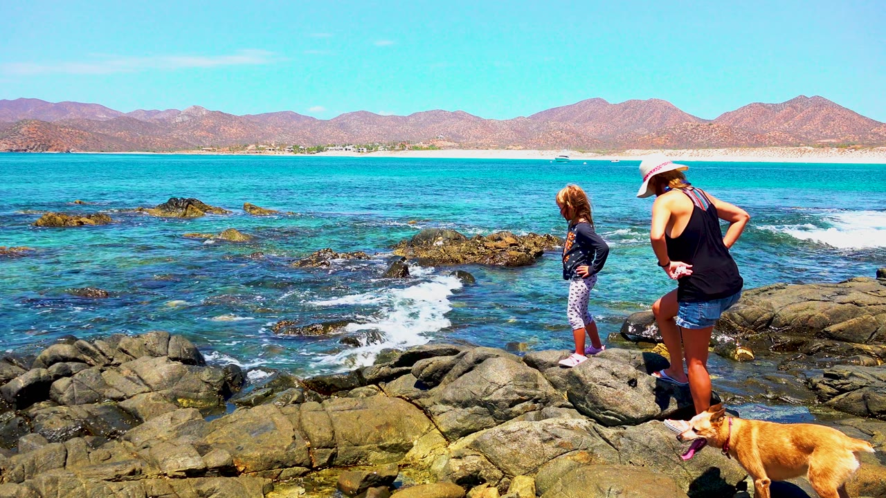 At 0:00 in El Sargento La Ventana, Mexico, a woman in a white hat, a child, and a brown dog stand on shoreline rocks beside turquoise water and distant mountains.
