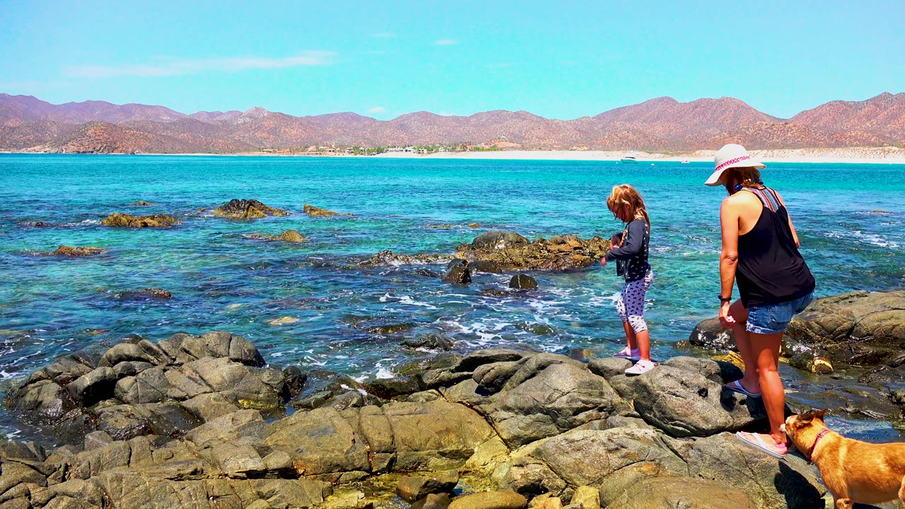 At 0:02 in El Sargento La Ventana, Mexico, the woman and child remain on the right side of the rocky shore as wave foam breaks around darker rocks in the water.