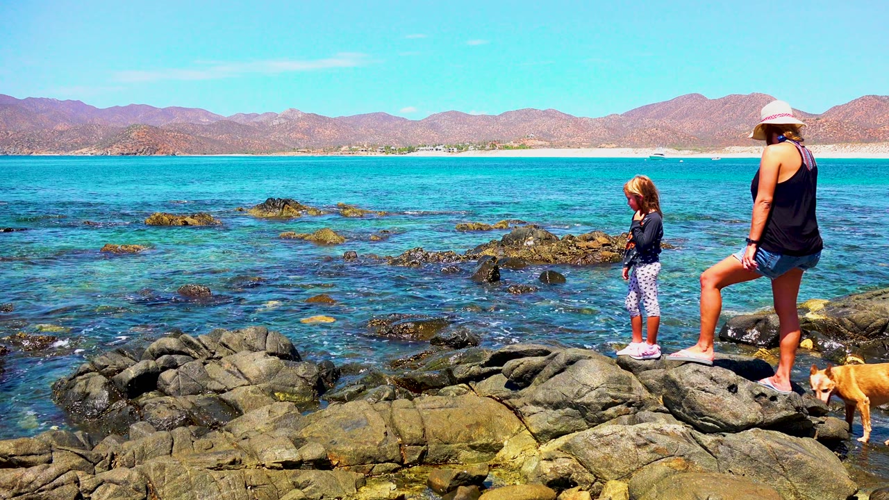 At El Sargento La Ventana Mexico, a woman in a sun hat stands on shoreline rocks while a child faces the water and a small dog appears at the lower right edge.