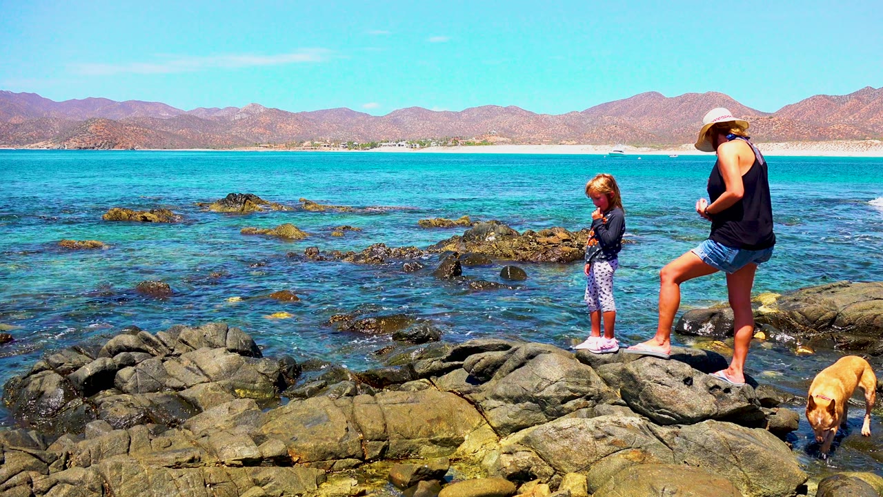 At El Sargento La Ventana Mexico, the woman stands with one foot raised on the rocks, the child looks downward near the water, and the dog is partly visible at the lower right.