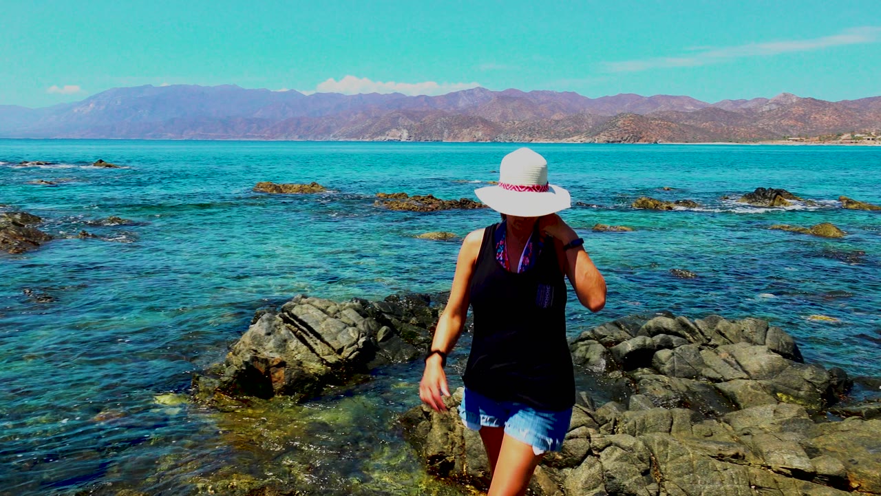 At El Sargento La Ventana Mexico, a woman in a white sun hat walks across dark shoreline rocks with clear turquoise water and distant mountains behind her.